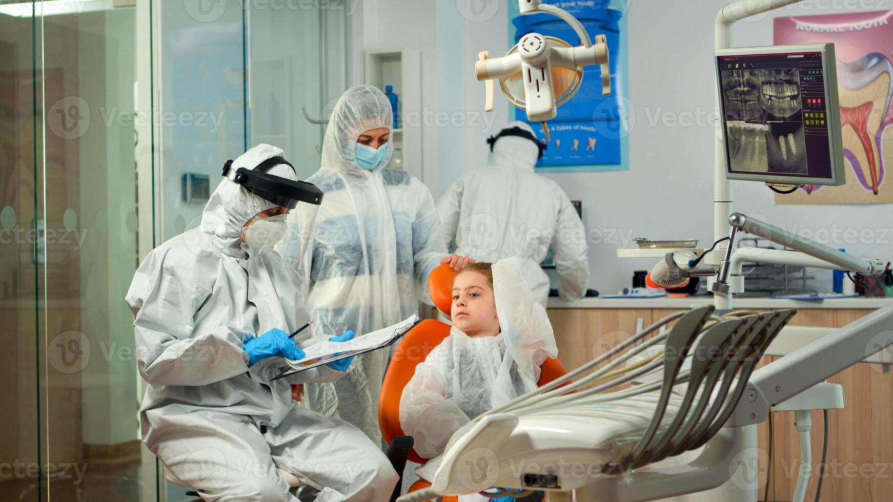 Pediatric dentist and nurse in ppe suit interrogating kid patient and taking notes on clipboard before examining. Stomatologist and assistent working in new normal dental office wearing coverall photo