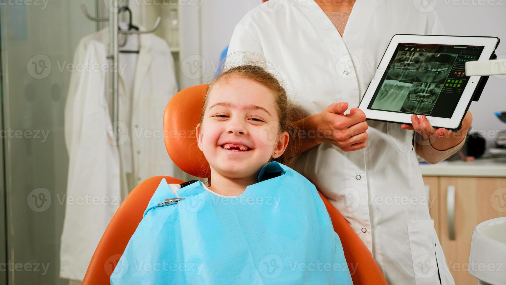 Close up of happy girl patient looking at camera laughing, waiting for pediatric stomatologist in dental unit. Kid lying on stomatological chair smiling at webcam while doctor talking with mother photo