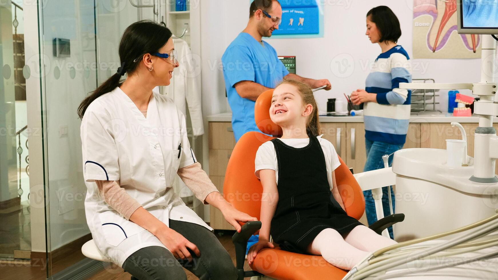 Little girl patient waiting pediatric stomatologist woman sitting in dental chair while man assistant talking with mother in background. Dentist discussing with girl before examining oral health. photo