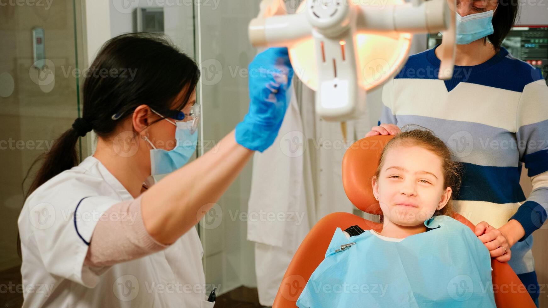 Woman stomatologist technician lighting the lamp for examining little patient sitting on stomatological chair. Doctor speaking to girl checking teeth health while nurse preparing tools for examination photo