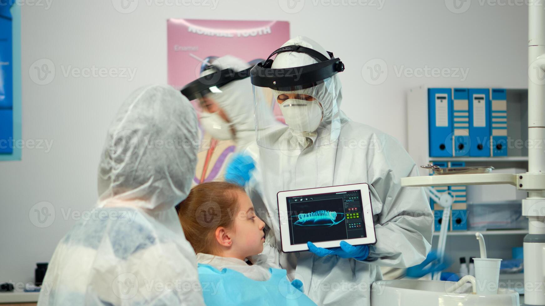 Dentist with ppe suit pointing on digital screen explaining x-ray to mother of girl patient. Medical team and patients wearing face shield coverall, mask, gloves, showing radiography using notebook photo