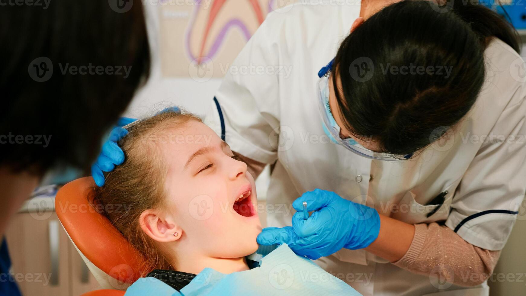 Close up of little patient lying on chair with open mouth during dental examination. Stomatologist holding sterilized tools talking with girl before stomatological intervation. Doctor wearing mask photo