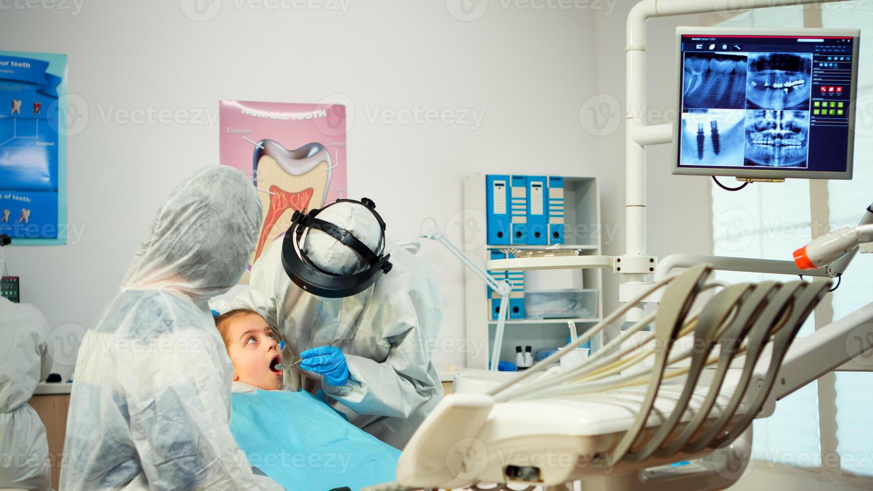Dentist in ppe suit standing near girl patient talking with mother, examining kid in new normal stomatological office. Concept of doctor visit in coronavirus outbreak wearing coverall and face shield photo