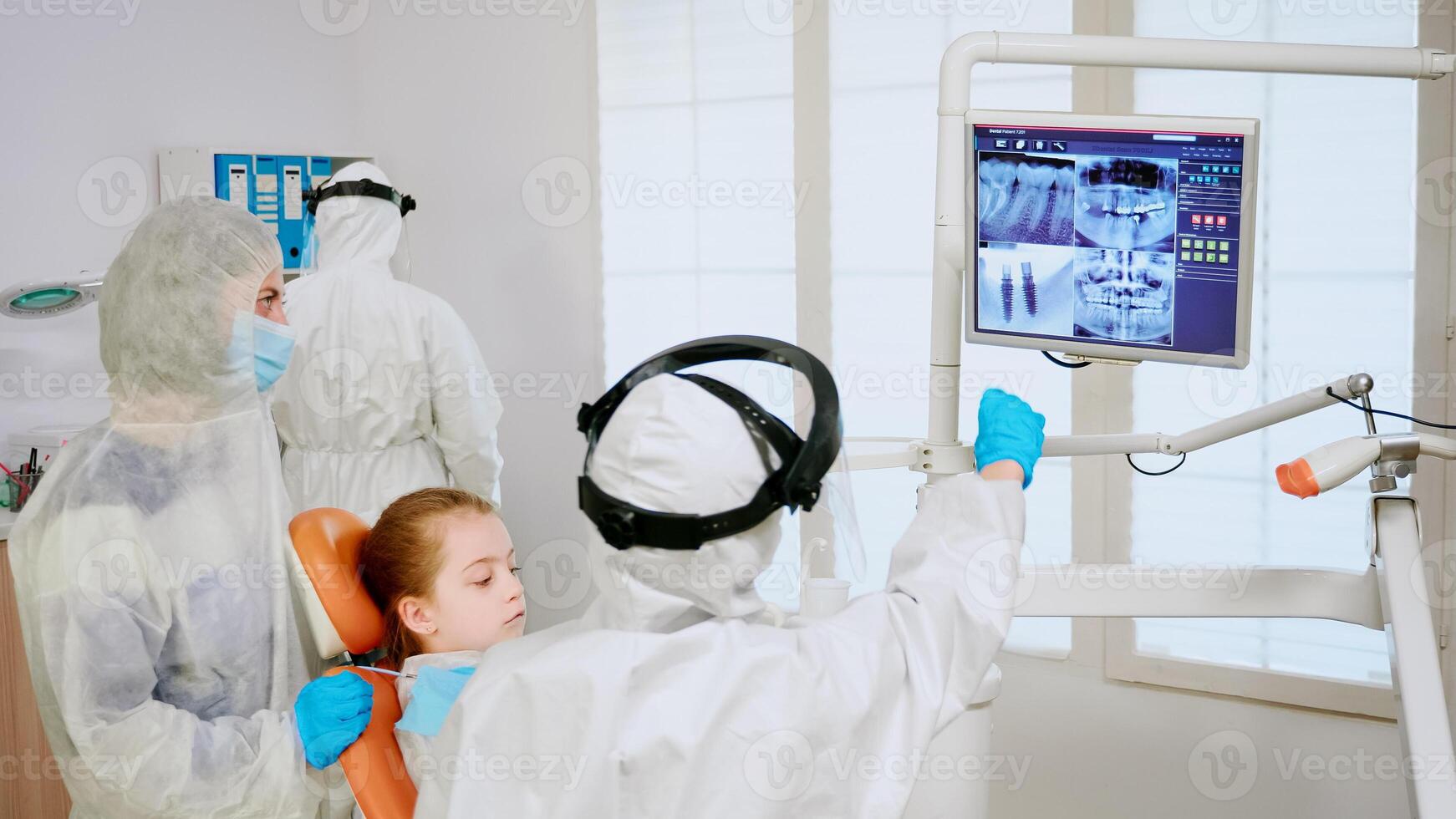 Stomatologist in coverall explaining dental problem using digital monitor during global pandemic. Assistant and doctor with coverall face shield mask gloves examining woman from stomatological chair photo