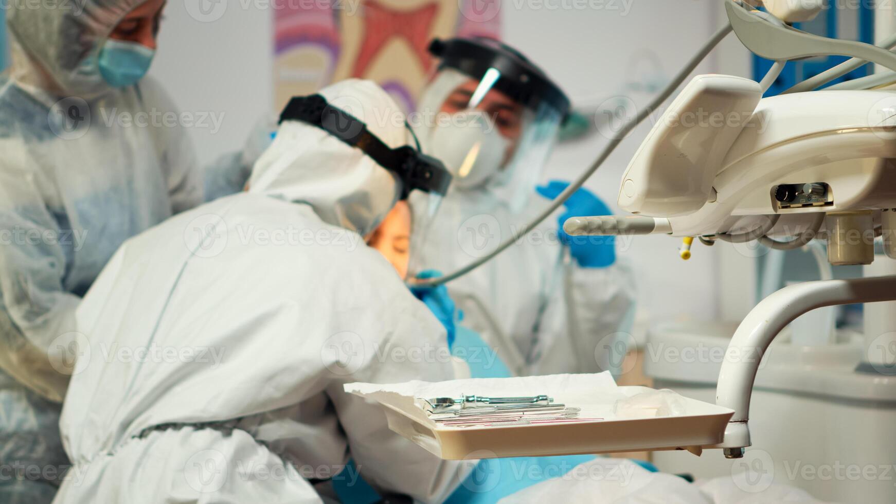 Close up of dentistry doctor in coverall using drilling machine for examining kid patient during global pandemic. Medical team wearing ppe suit, face shield, mask, gloves in stomatological office photo