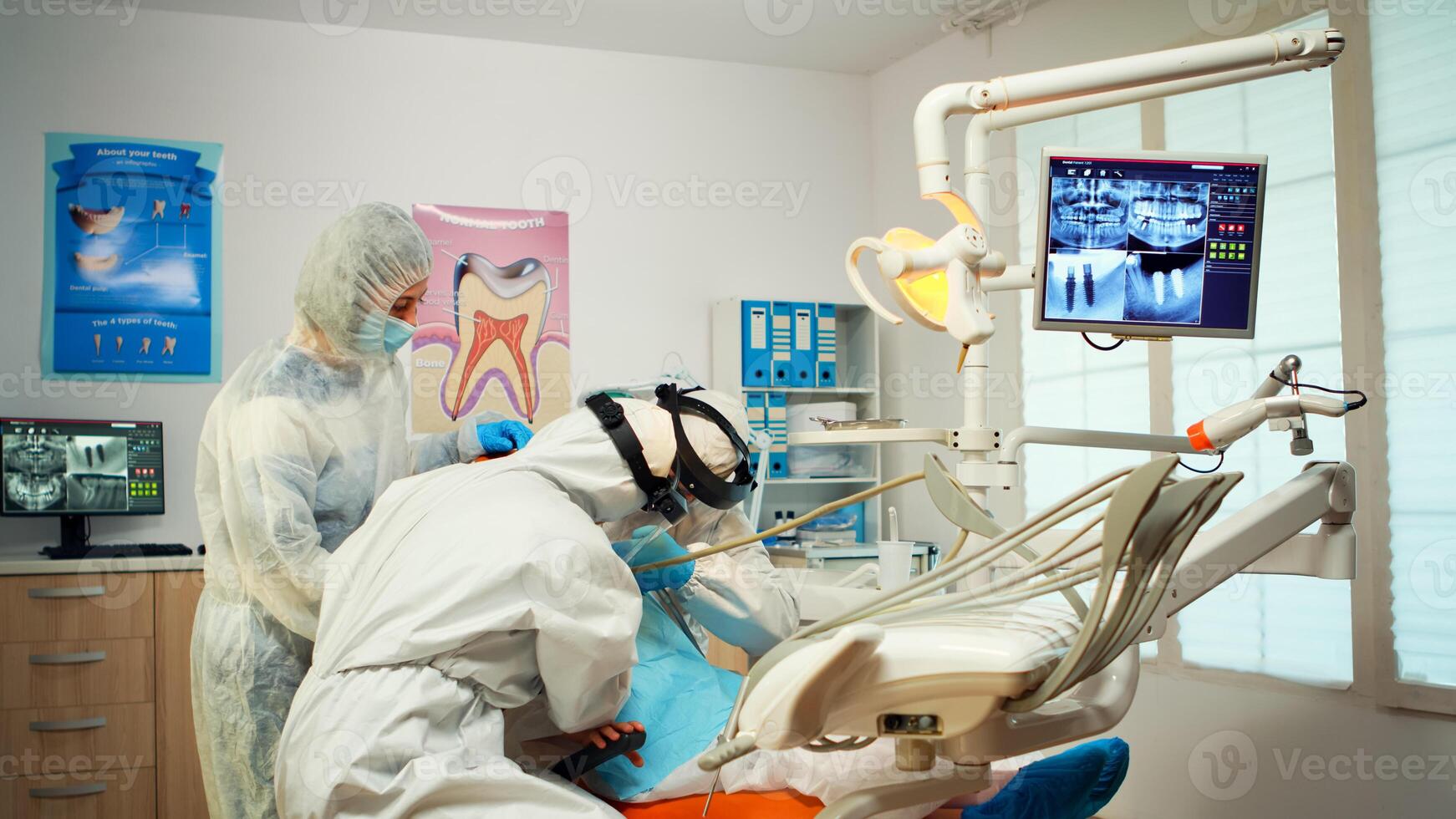 Dental technician in protective equipment lighting the lamp for examining child patient during covid-19 epidemic. Medical team talking with mother wearing face shield, coverall suit, mask and gloves photo
