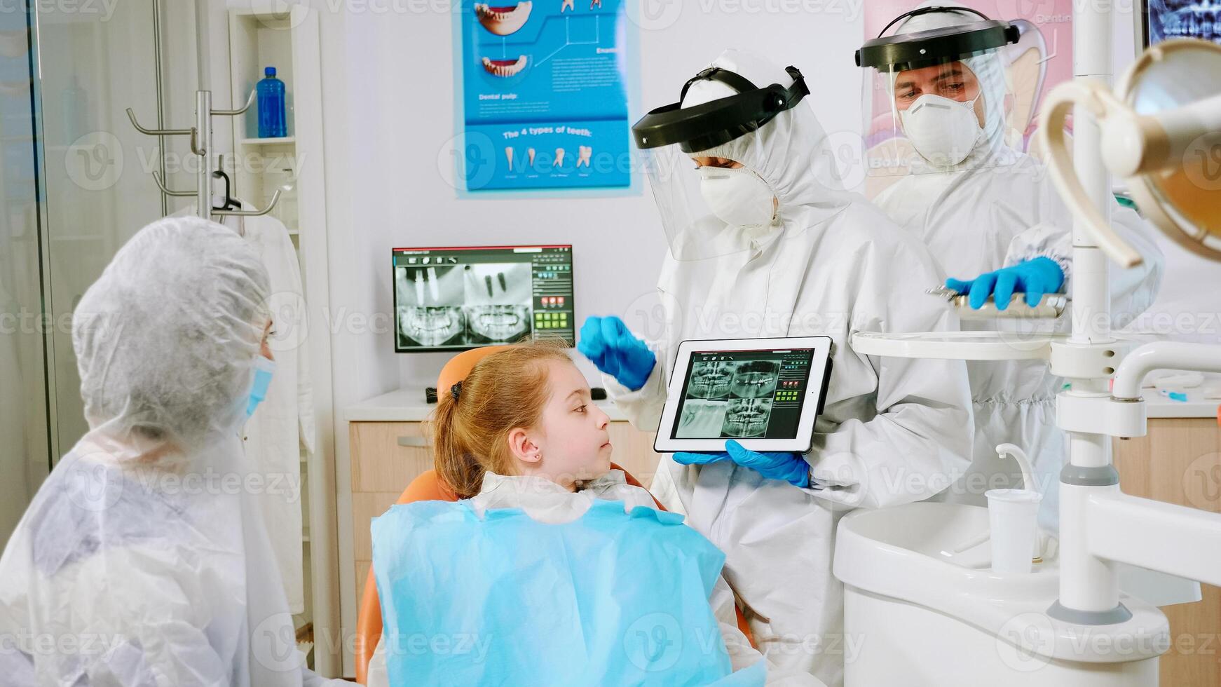 Stomatologist in protective equipment showing on tablet dental x-ray reviewing it with mother of patient. Medical team wearing face shield mask, gloves, explaining radiography using notebook display photo