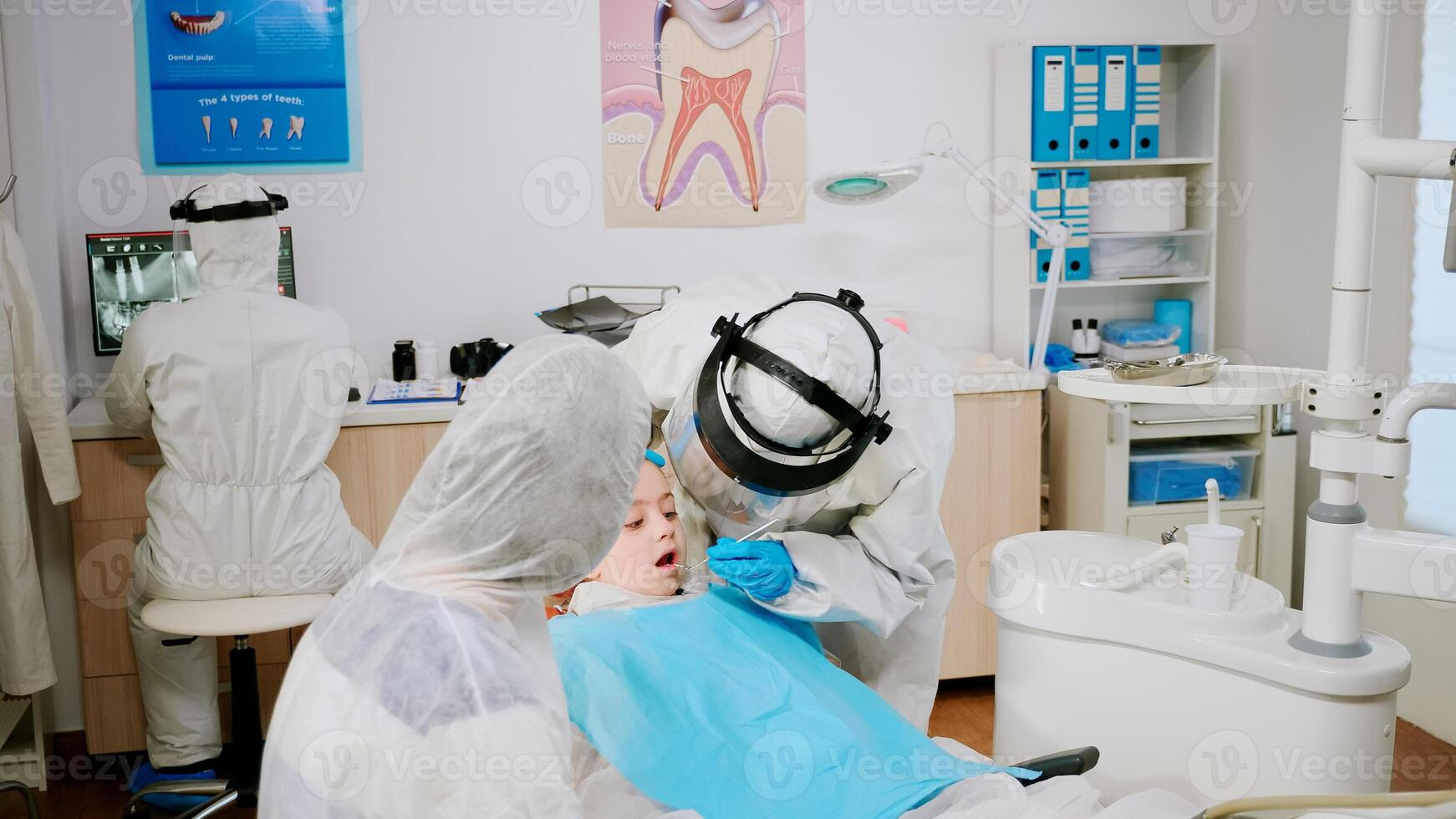 Pediatric dentist in protective shield checking teeth of little girl sitting in stomatological chair during coronavirus pandemic. Doctor wearing face shield, protection suit, mask and gloves. photo