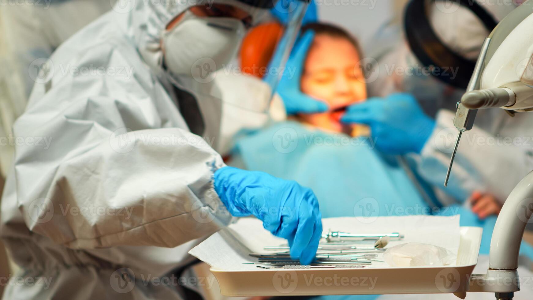 Medical team working with sterilized tools wearing face shield, coverall suit, mask and gloves during coronavirus outbreak in new normal stomatological clinic. Dentist treating teeth of little girl photo