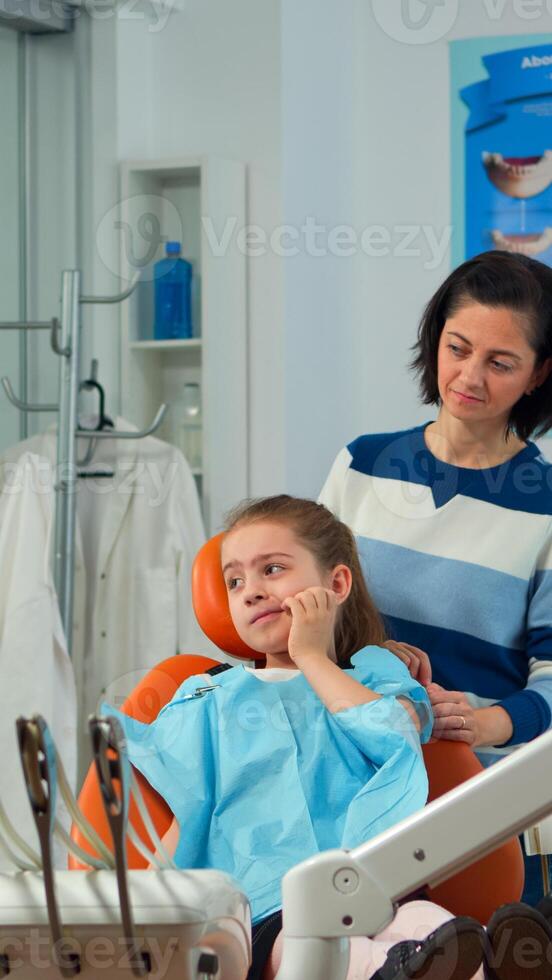 Pediatric woman dentist talking with mother and girl about eating habit and oral hygiene of the child before dental examination. Sad kid patient is showing sick tooth while nurse preparing dental tools photo