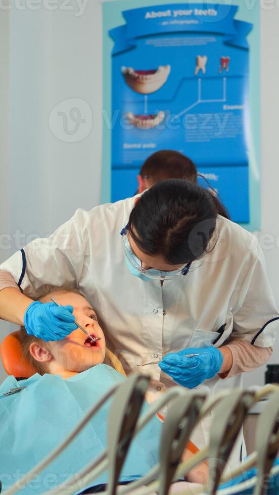 Stomatologist talking with mother of kids, lighting the lamp and examining little girl standing near stomatological chair. Pediatric dentist speaking to woman while nurse preparing sterilized tools. photo