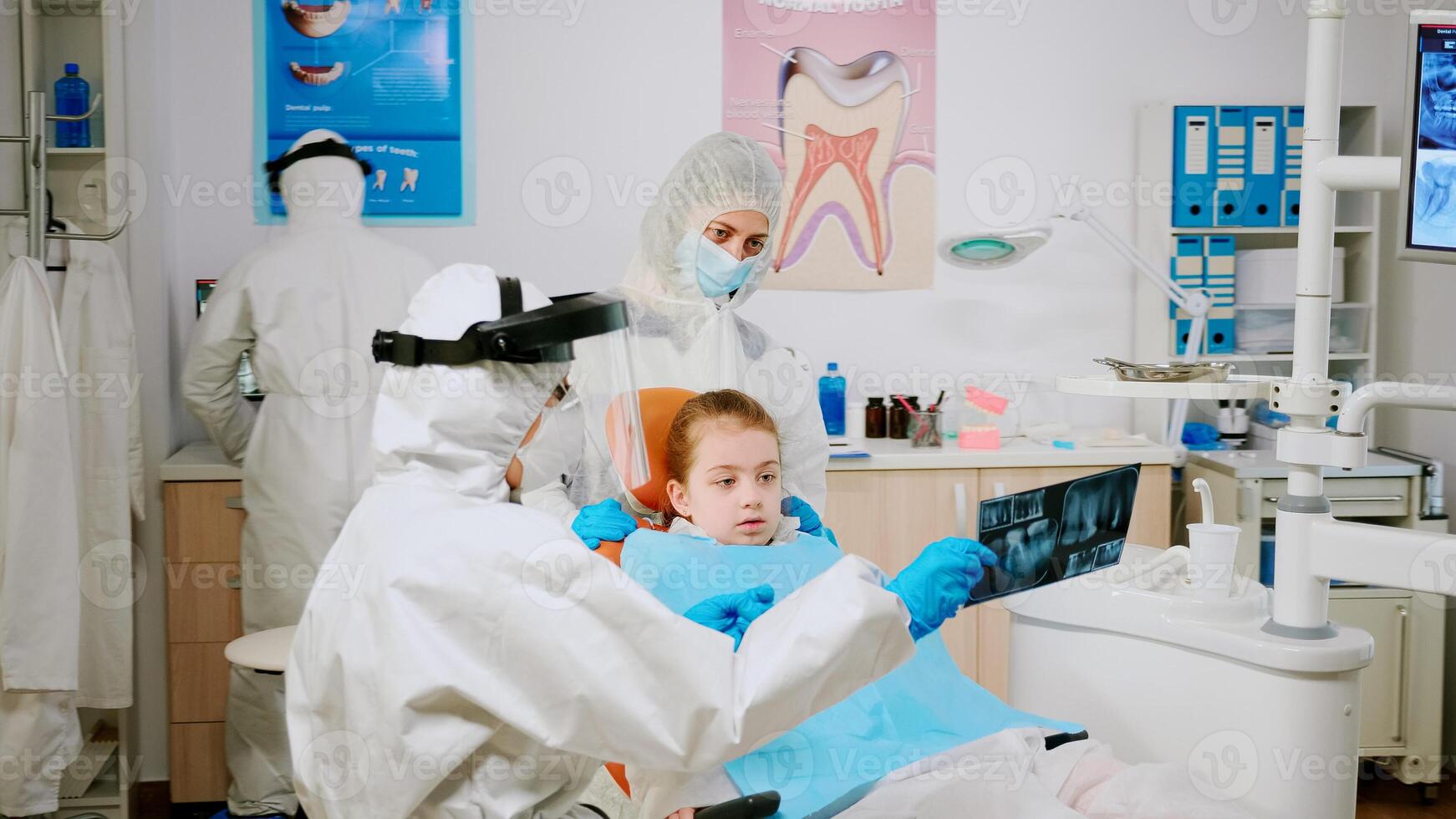 Doctor in protective suit discussing about x-ray of tooth with mother of child patient explaining treatment using tablet in covisd-19 pandemic. Medical team wearing face shield, coverall, mask, gloves photo