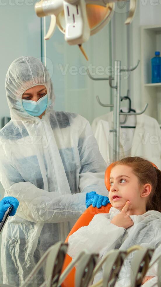 Dentist doctor in ppe suit interrogating kid patient and taking notes on clipboard while girl indicating affected mass. Stomatologist and assistent working in new normal dental office wearing coverall photo
