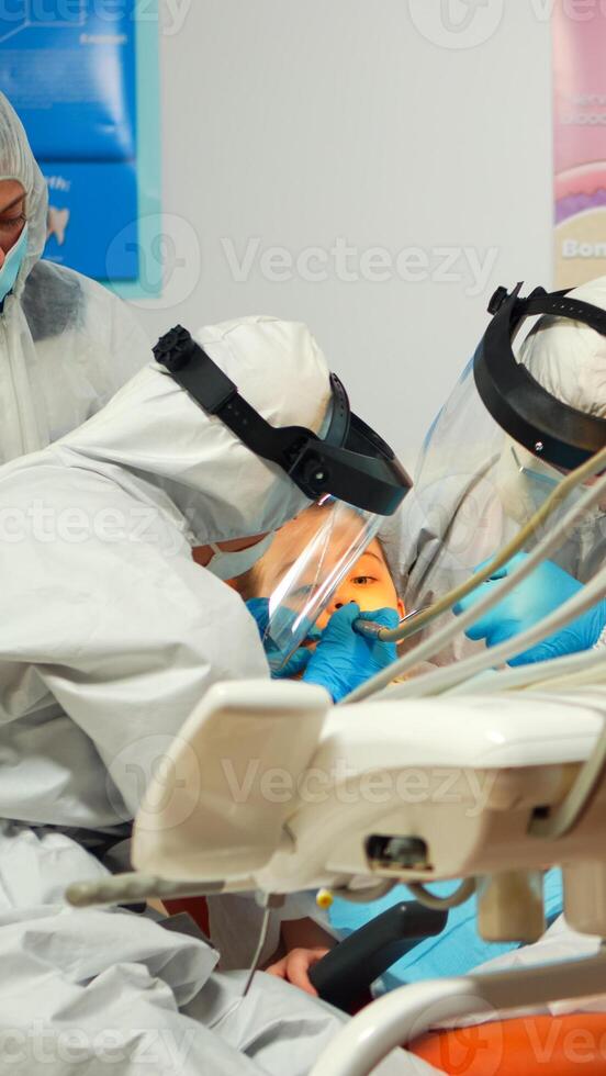 Dentist technician in coverall drilling kid tooth in dental clinic during coronavirus pandemic. Concept of new normal orthodontic visit in coronavirus outbreak wearing protective suit and face shield photo