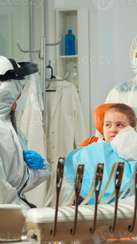 Child in protection suit showing dental problem to orthodontist wearing coverall during global pandemic. Concept of new normal dentist visit in coronavirus outbreak wearing face shield photo