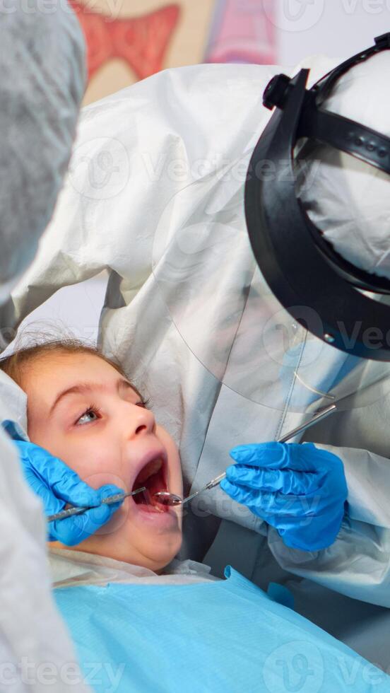 Close up of dentist in coverall examining girl patient in new normal stomatological office, standing near stomatological chair. Concept of doctor visit in coronavirus outbreak wearing face shield photo