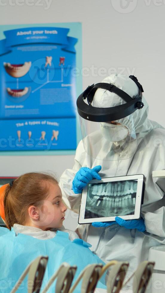 Dentist in protective equipment showing on tablet dental x-ray reviewing it with mother of kid patient. Medical team wearing face shield coverall, mask, gloves, explaining radiography using notebook photo