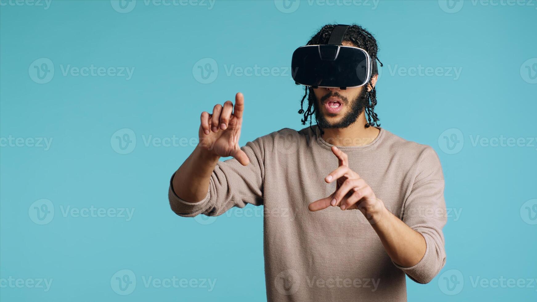 Tech enthusiast wearing virtual reality goggles, doing swiping gestures. BIPOC man using high tech futuristic modern VR glasses, making tactile touching gesturing, blue studio background photo