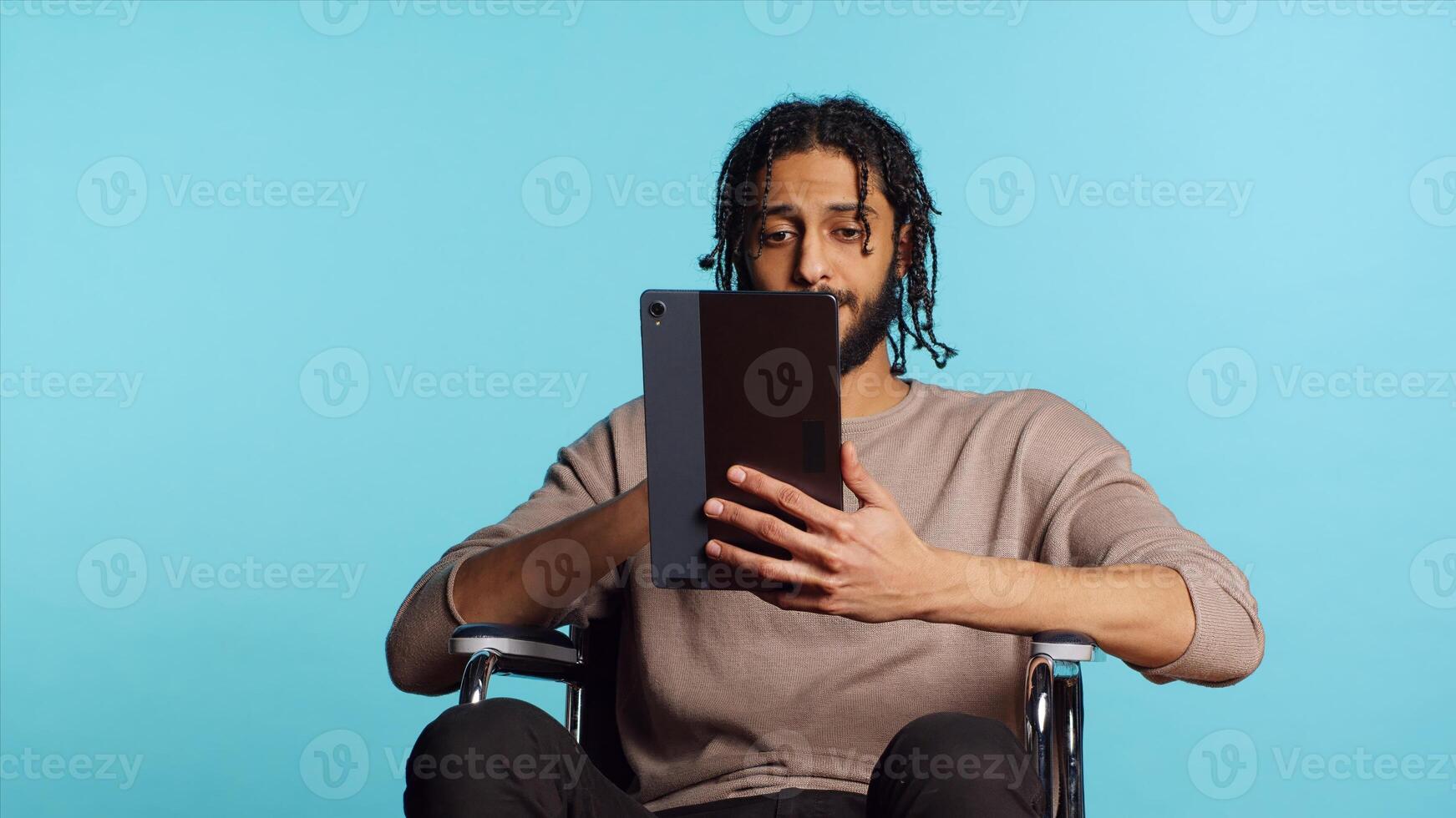Person with paraplegia using tablet to post images on social media, doing selfie and writing photo caption. Man in wheelchair using online editing application on digital device, studio, camera A