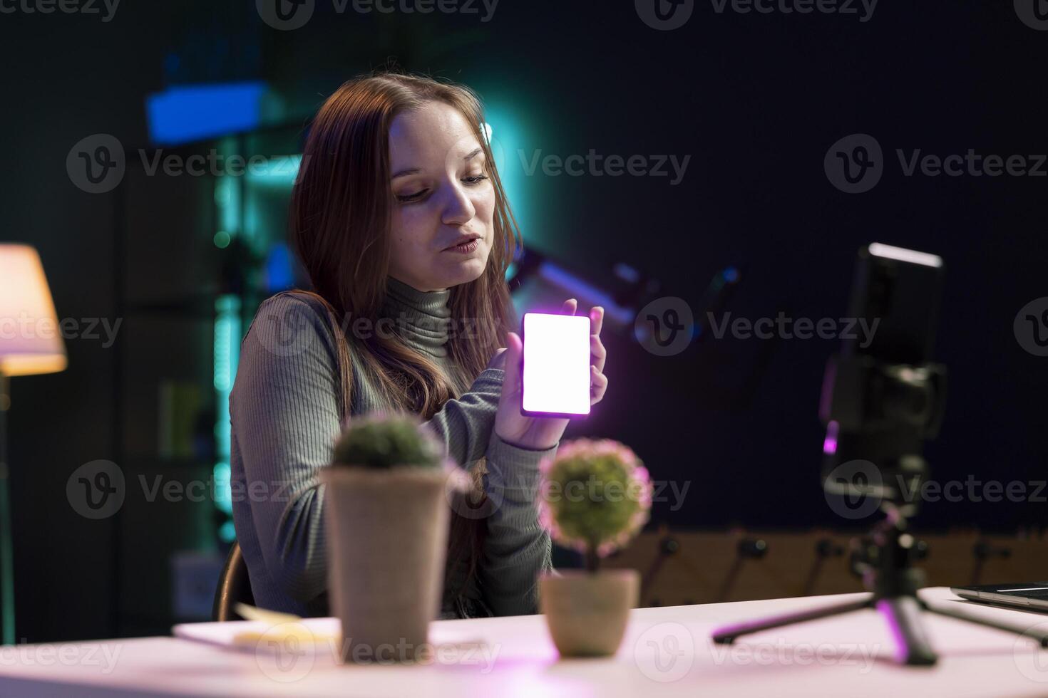 Gen Z girl in apartment studio using mobile phone to record, reviewing mini RGB lights. Young entertainer films electronics haul with cellphone on tripod, presenting LED lighting device photo