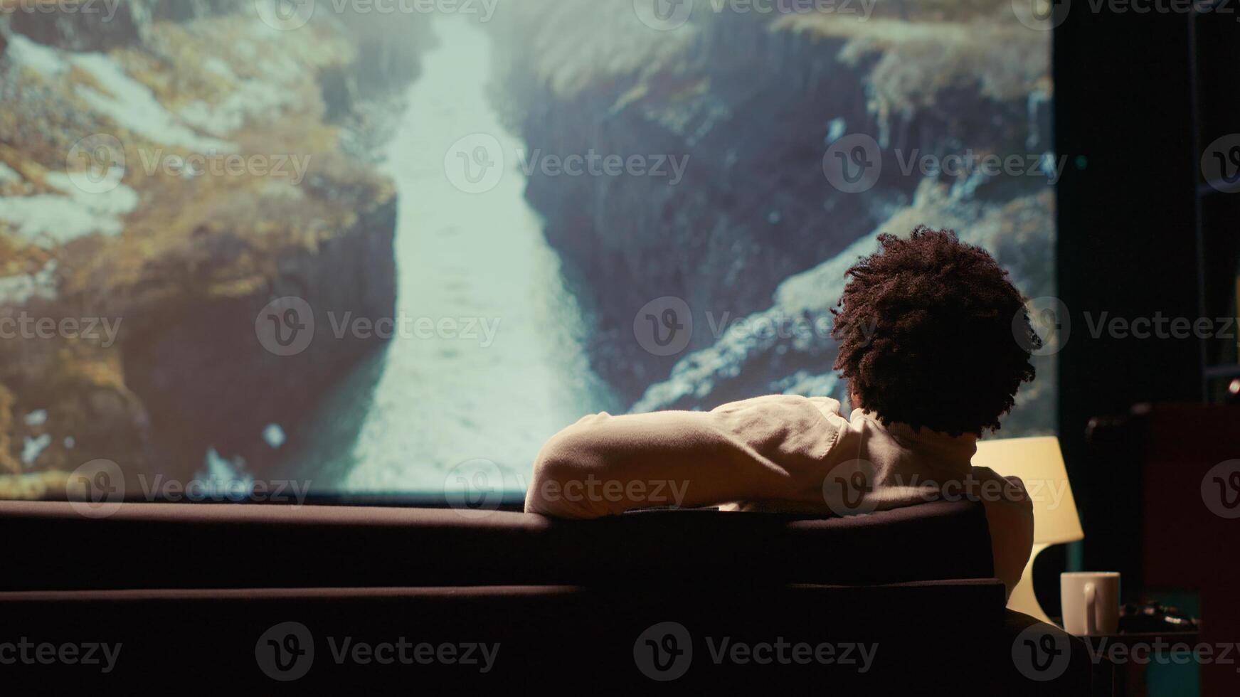African american man watching nature documentary with breathtaking cinematography on huge TV screen. Person enjoying scenery shots on widescreen television set, relaxing on couch photo