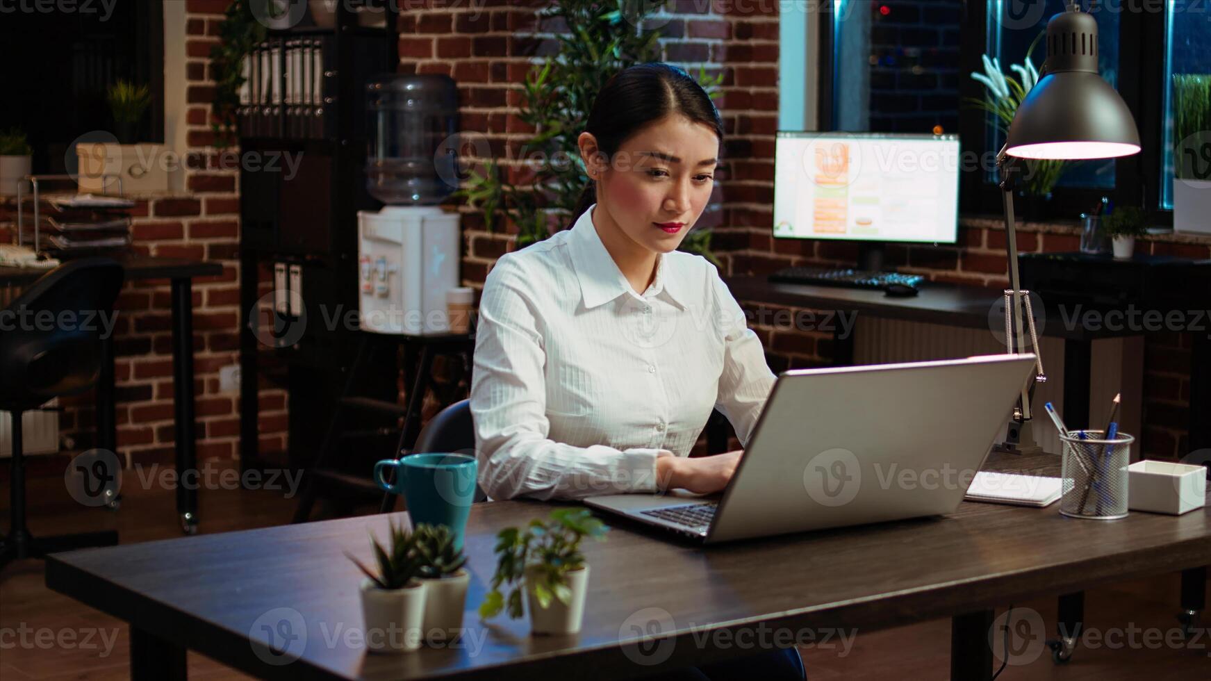 Smiling woman sitting down at office desk, typing on laptop keyboard, starting work with mug full of coffee, solving tasks late at night. Cheerful employee imputing data on computer photo