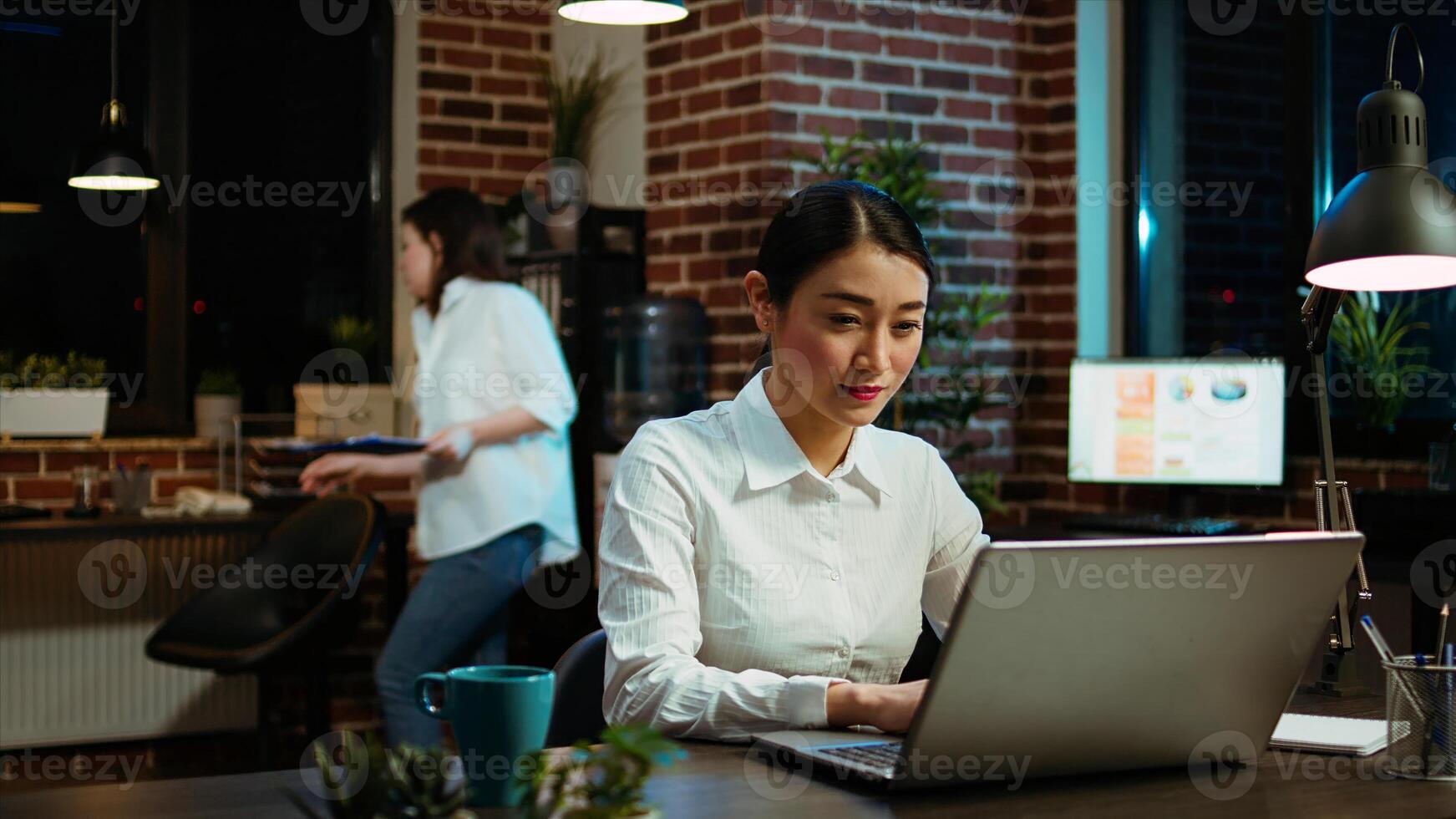 Portrait of smiling asian businesswoman doing computer tasks for team project in office. Cheerful employee working late at night, typing on laptop keyboard next to coworker, camera A photo