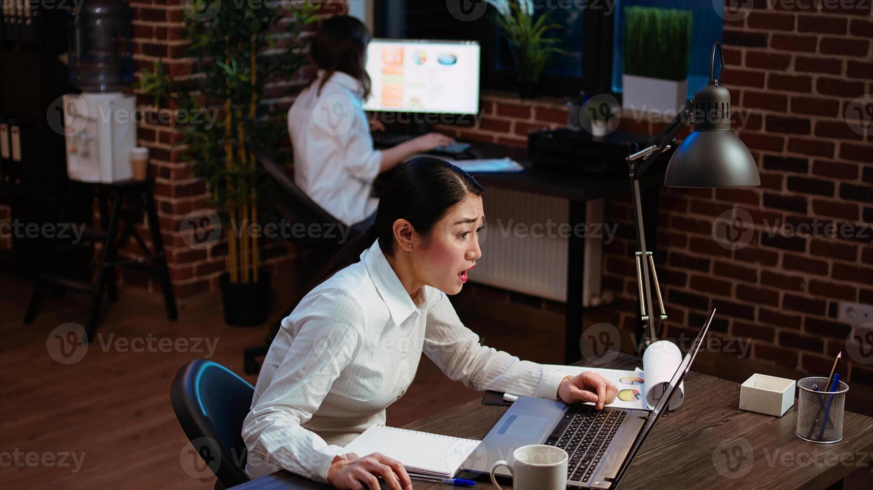 Bookkeeper feeling dread while having to do complicated tax reports in office at night. Worker squinting eyes in exasperation, feeling overworked while using laptop to do project research, camera A photo