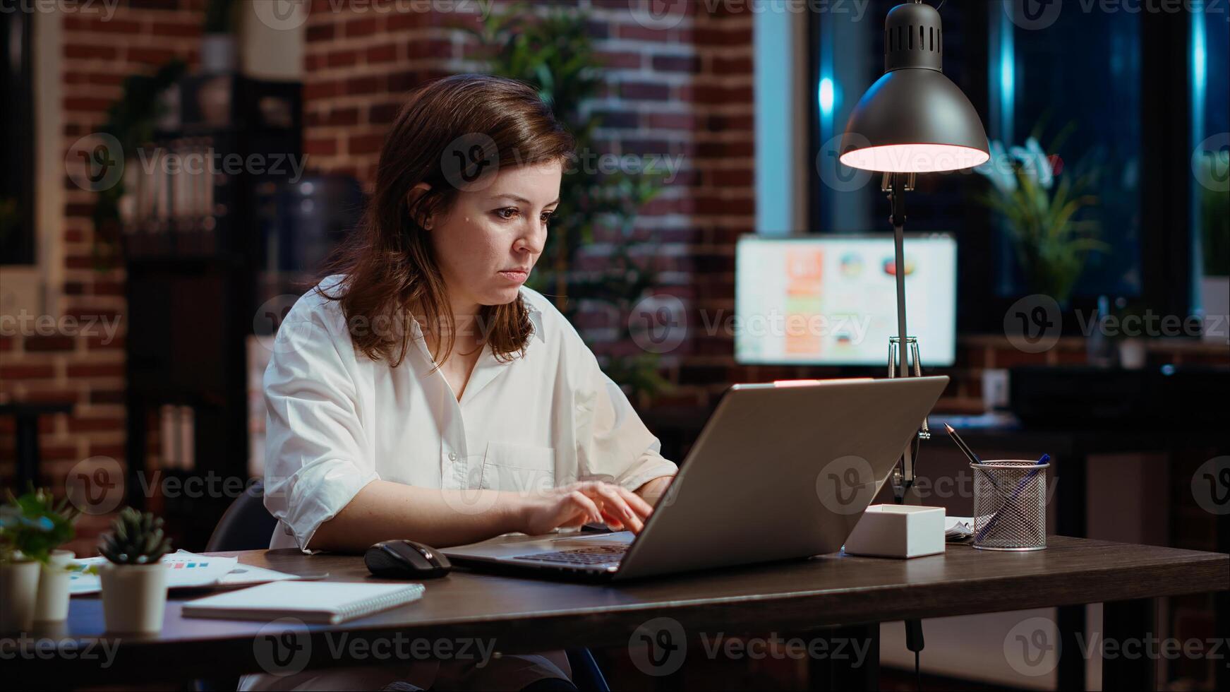 Office clerk looking through financial business documents on laptop late at night in workplace. Employee working on company project with deadline approaching soon during nightshift photo