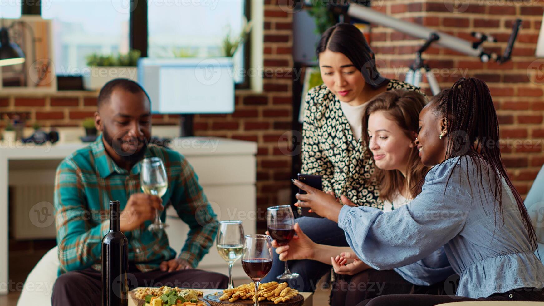 Multiracial friends at apartment party looking at social media posts made by acquaintance on cellphone, making fun of them. Amused guests in living room reacting to cringe images posted online by mate photo