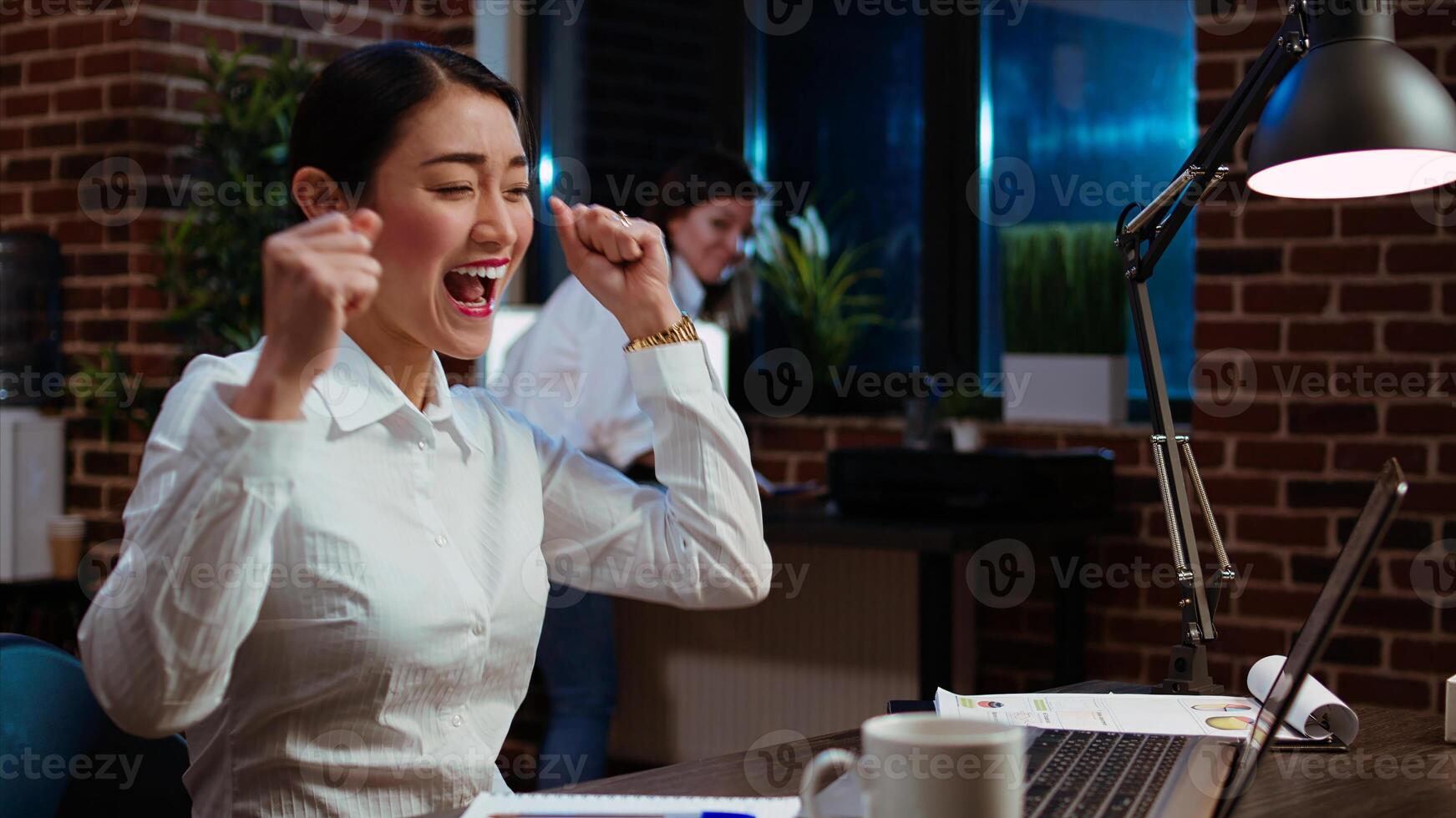 Happy office clerk celebrating finishing company project overnight by dancing on desk chair. Euphoric employee enthusiastic about finally finishing tasks on laptop, singing and jubilating photo