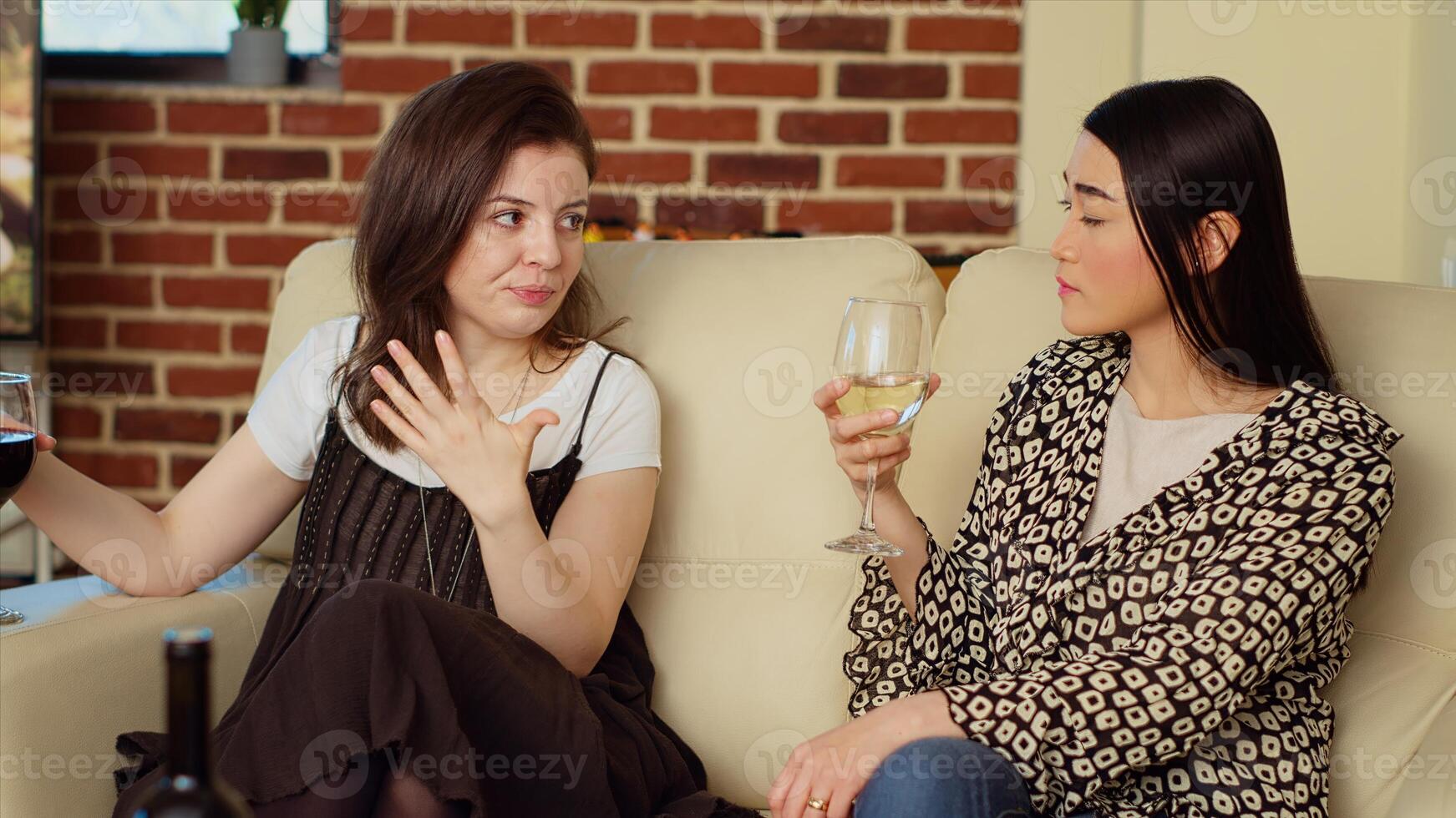 Asian woman chatting with friend on comfortable couch at apartment party, drinking wine and enjoying conversation. Joyful BFFs catching up with each other at home, consuming champagne photo