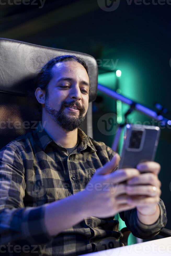 Cheerful male student is utilizing his mobile device to browse social media while seated at his desk. Bearded man with a smile answers texts while using his smartphone for surfing the internet. photo