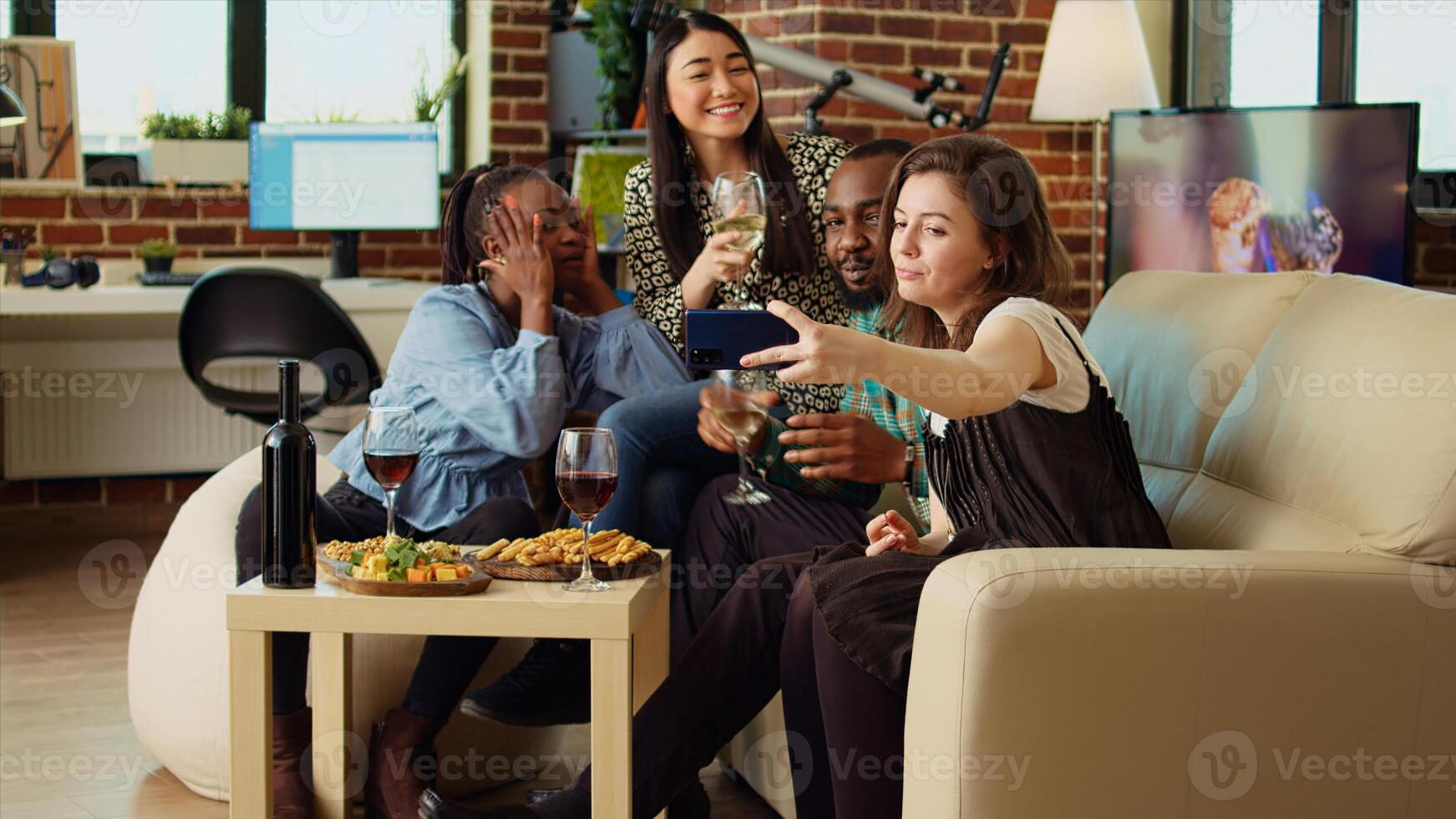 Multiethnic friends taking group pictures with cellphone at apartment gathering. Happy people in living room posing for photo, making funny faces and drinking alcoholic beverages photo