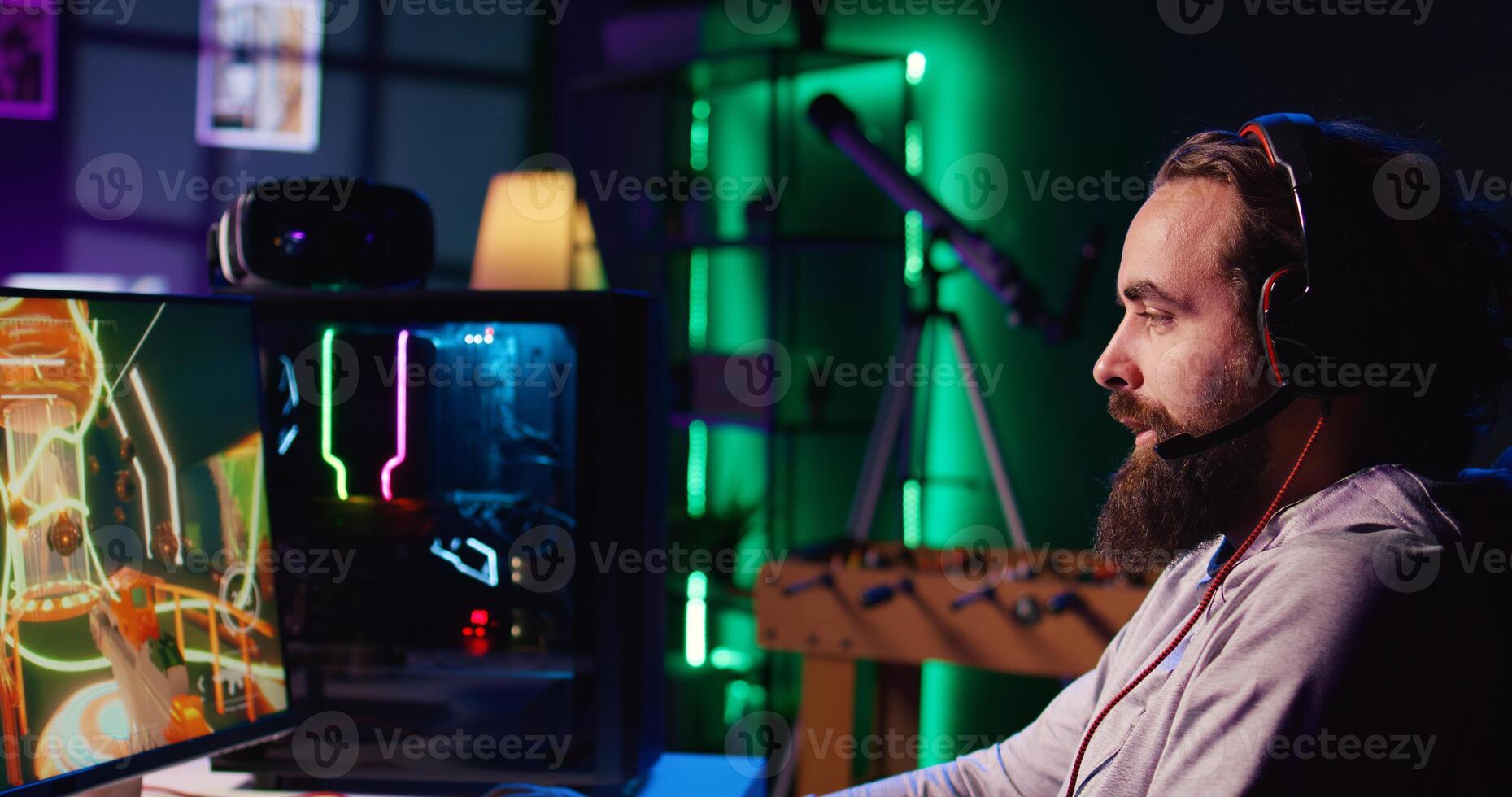 Happy man using headphones mic to communicate with online friends from his dark living room, playing engaging games on gaming PC at computer desk, relaxing after work photo