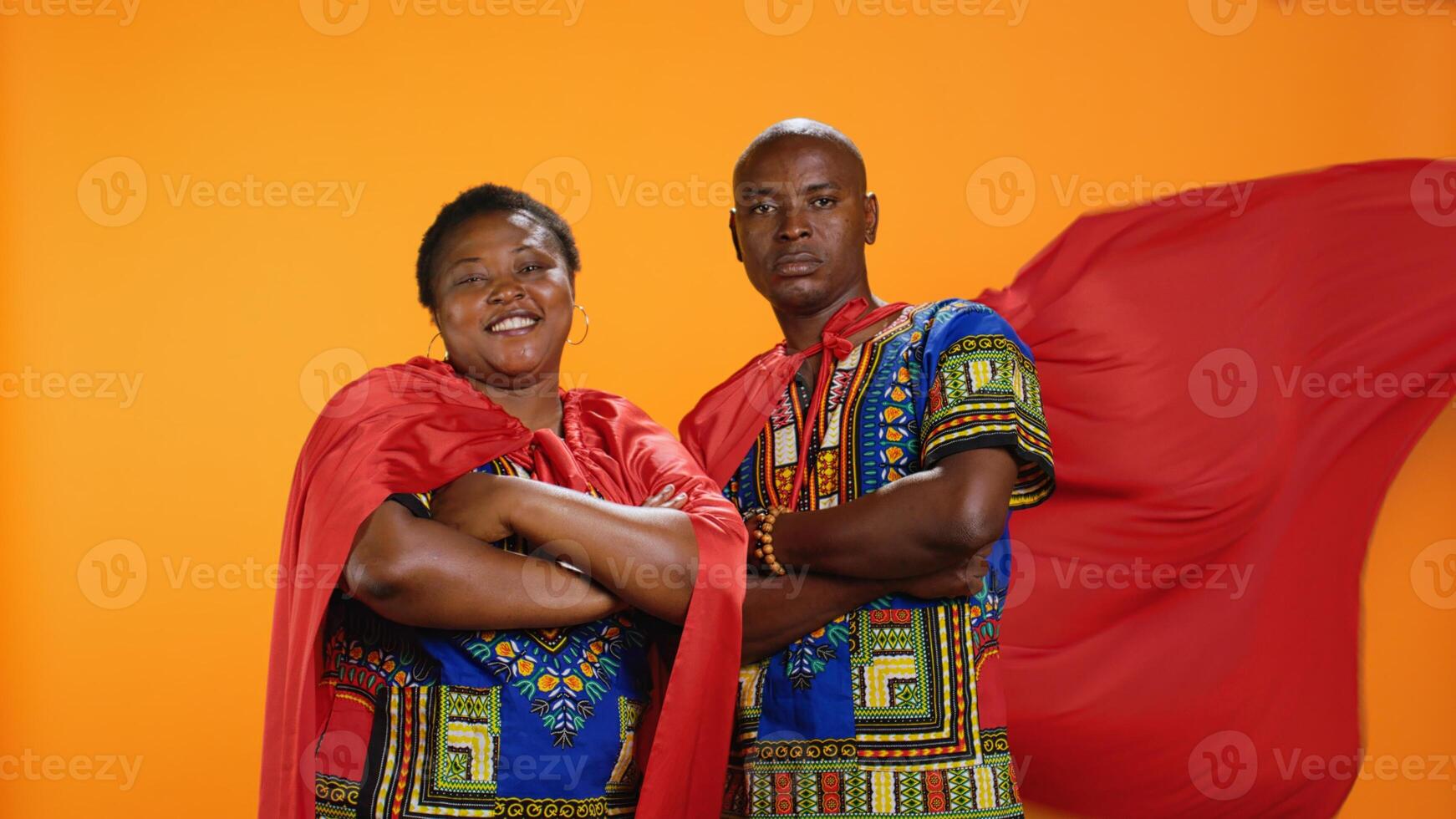 Ethnic married couple in superhero red capes posing on camera, feeling proud about their abilities over orange background. African american people acting as comic book heroes with cloaks. photo