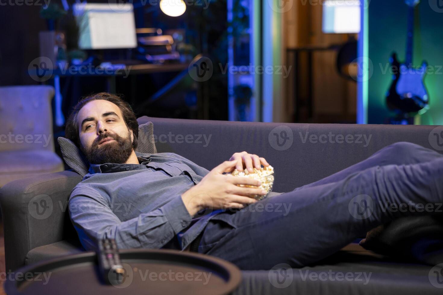 Spectator lounged on couch eating popcorn while watching interesting movie on streaming service, enjoying time off from work. Man laying down in apartment consuming snacks, looking at films photo
