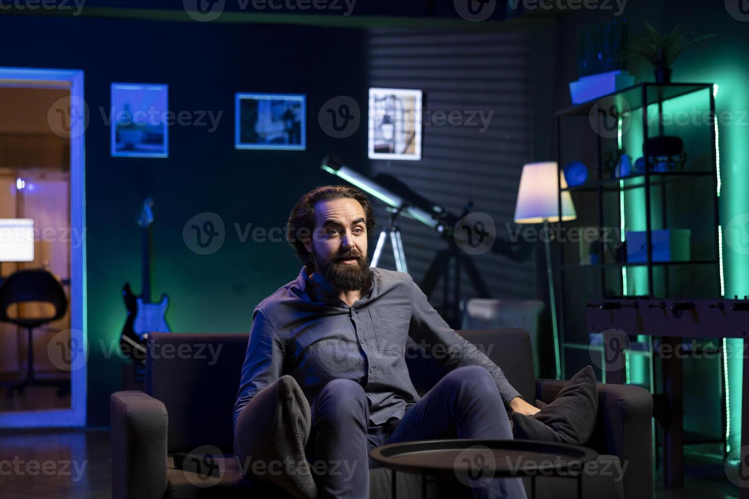 Man enjoying leisure time at home watching TV, amazed by plot twist. Viewer chilling on couch in neon illuminated apartment, entertained and astonished by television shows twisted turn of events photo