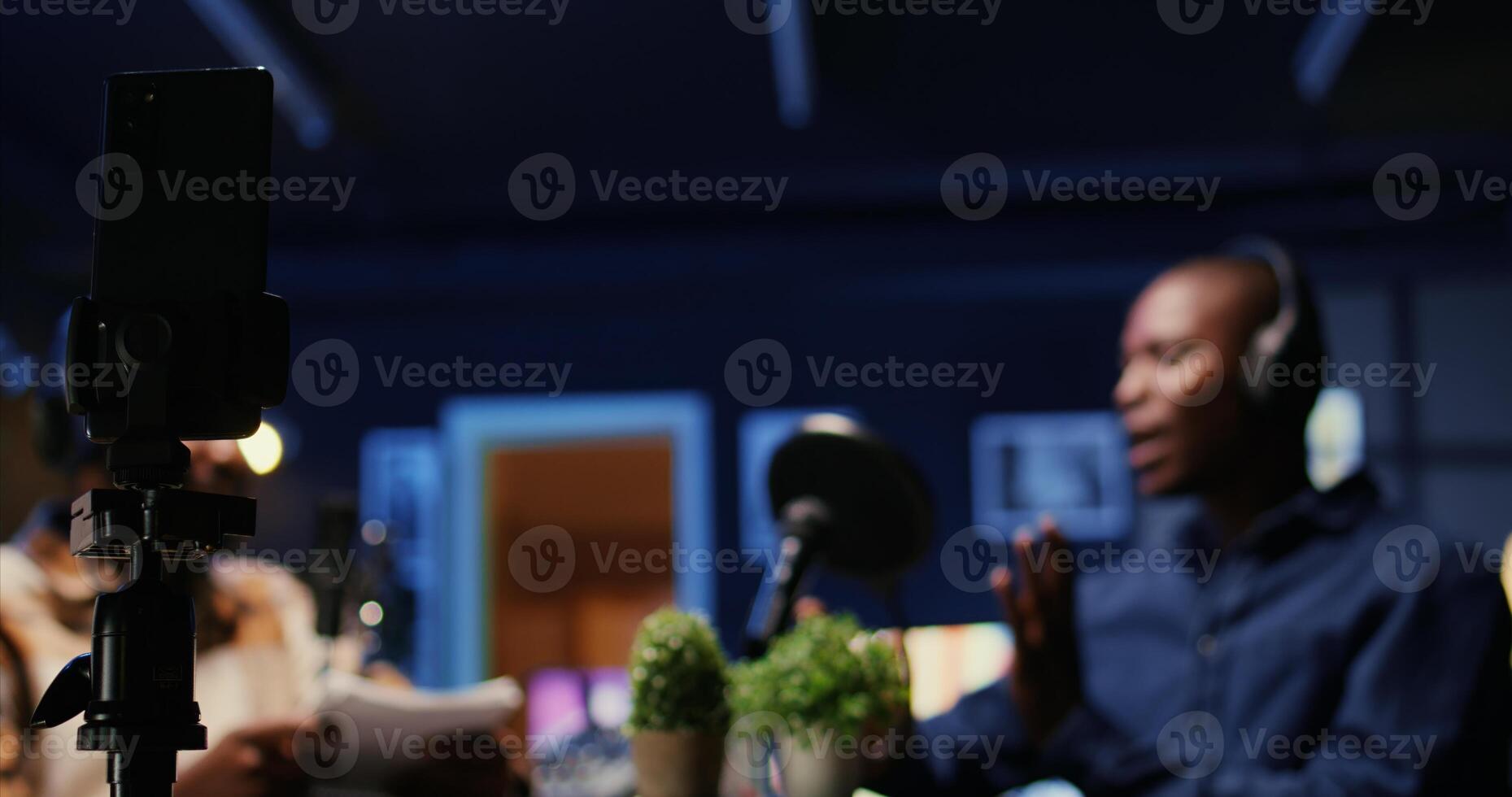 Focus on smartphone on tripod recording vlogger show host in blurry background interviewing guest in neon lights studio. Presenter records online podcast episode with mobile phone, close up photo