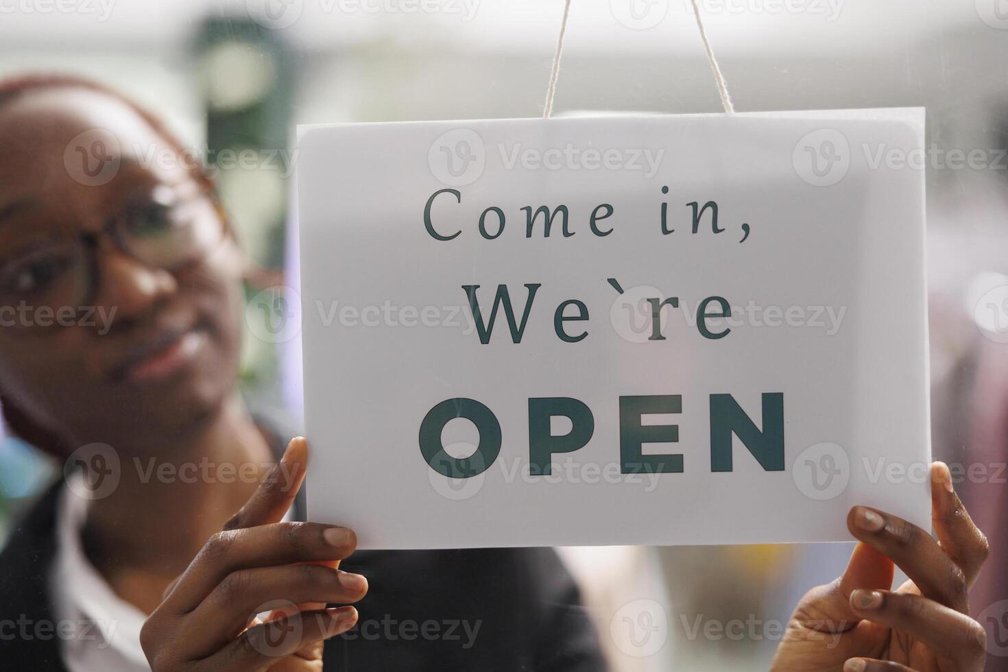 Fashion clothes shop manager holding opening signboard with come in message. Apparel store young african american woman assistant hanging sign informing clients about workday beginning photo