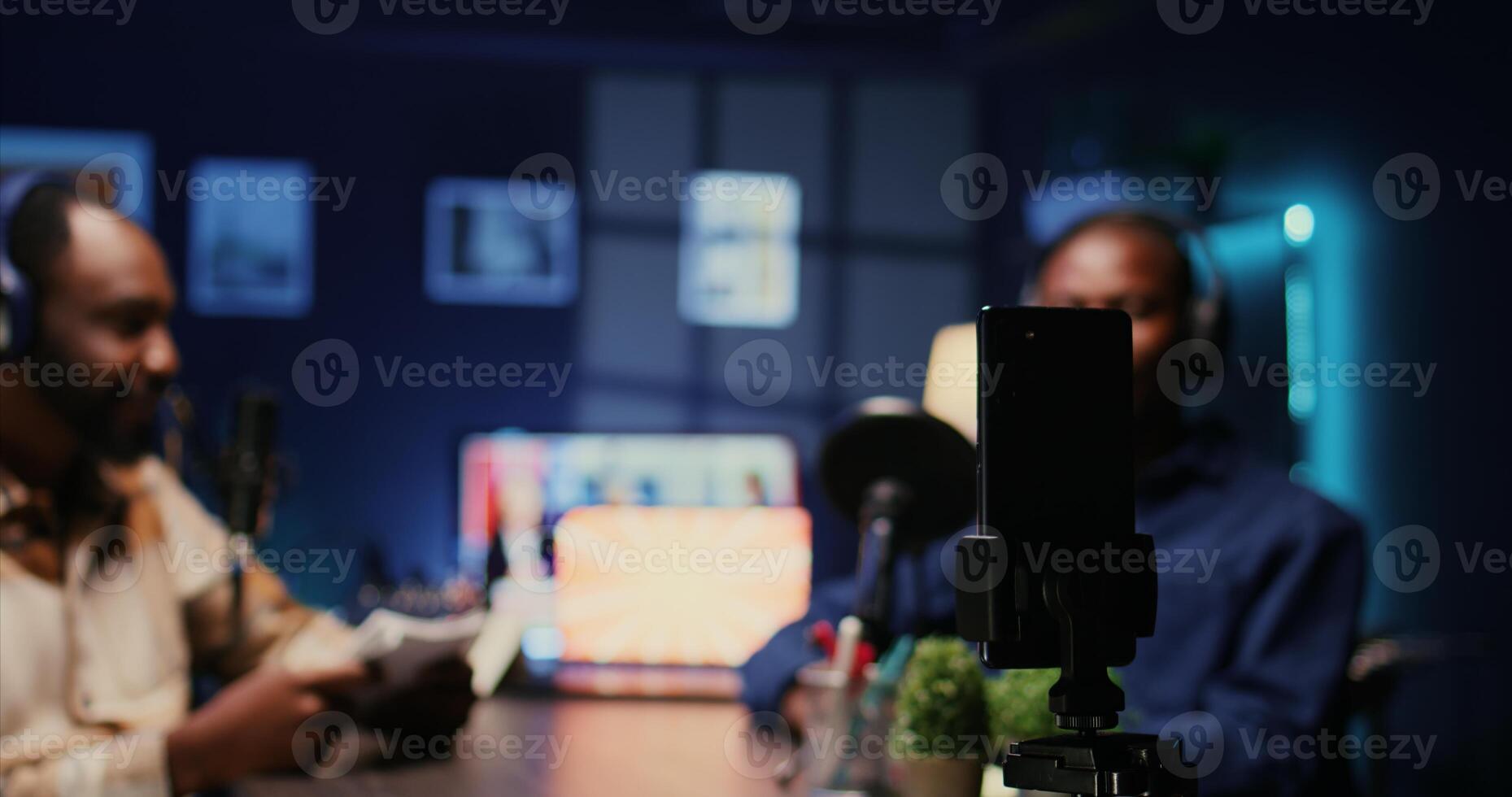 Focus on smartphone on tripod used by host in blurry background to record entertaining discussion during livestream in apartment studio. Close up shot of mobile phone used to produce comedy podcast photo