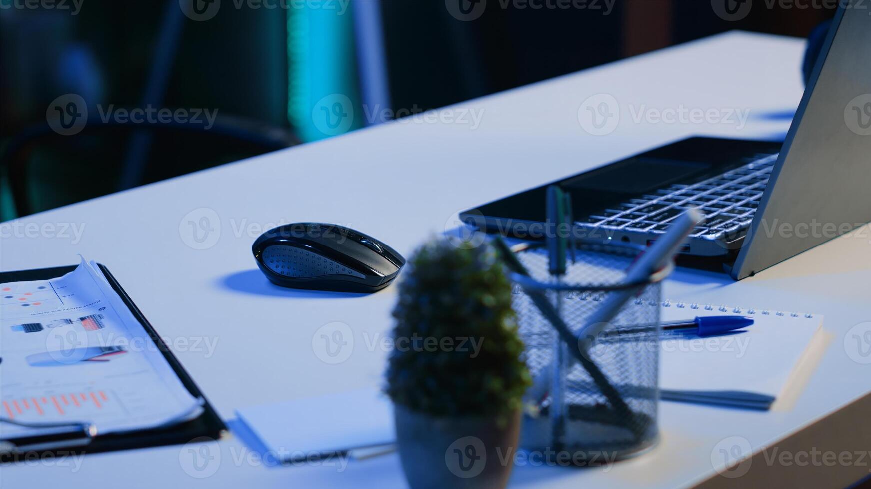 Office desk filled with productivity items in empty home interior illuminated with neon lights at night. Laptop on table and other work related objects in apartment room, camera A close up shot photo