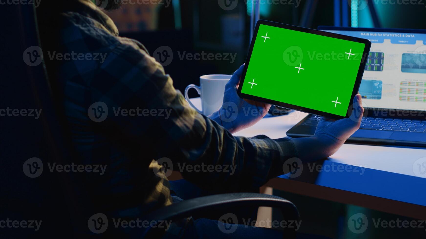 Man in home office checking statistical financial graphs on laptop, crosschecking with data on mockup tablet. Remote employee holding green screen device, analyzing business graphics photo