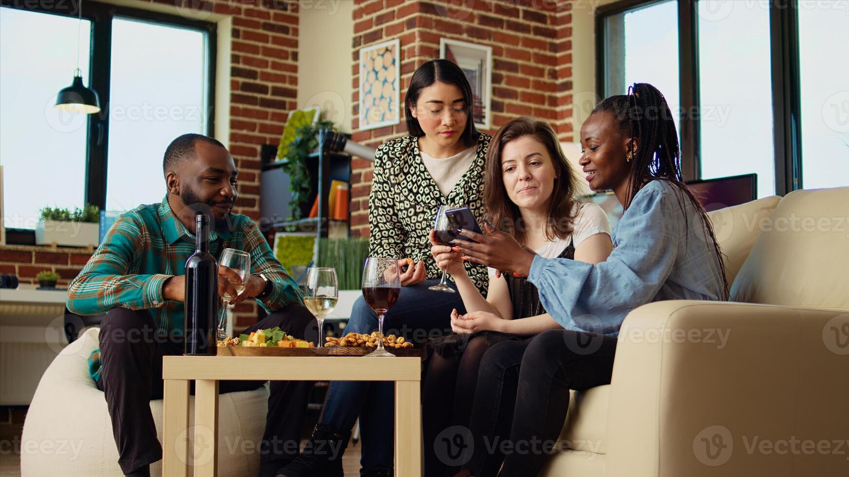 Friends at house party using smartphone to laugh at social media posts made by acquaintance while eating snacks and drinking wine. Guests using phone to look at images online, enjoying appetizers photo