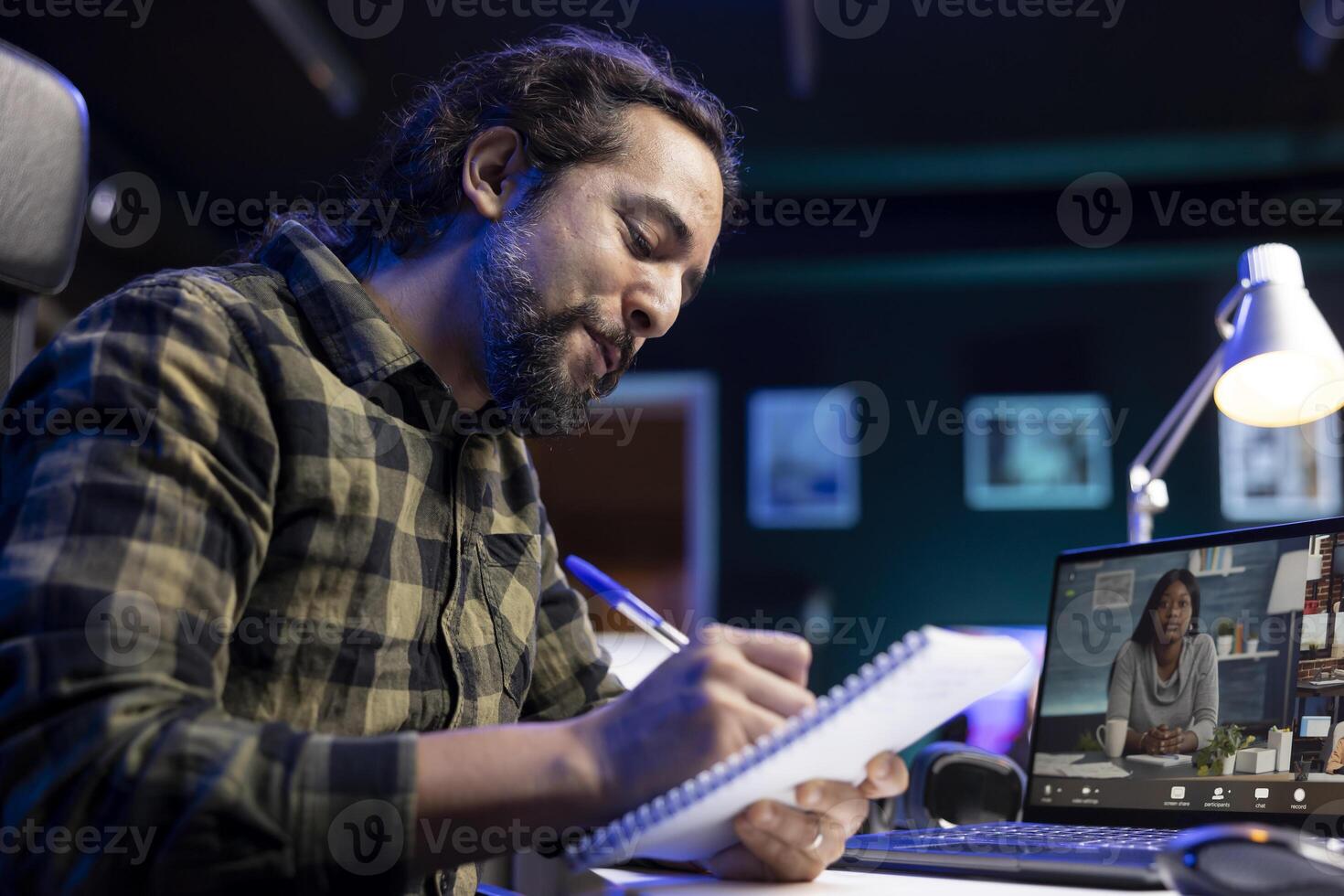 Closeup of bearded man with a pen and notebook, writing meeting notes while on call on the laptop. Male individual positively working from home, using his notepad and portable computer. photo