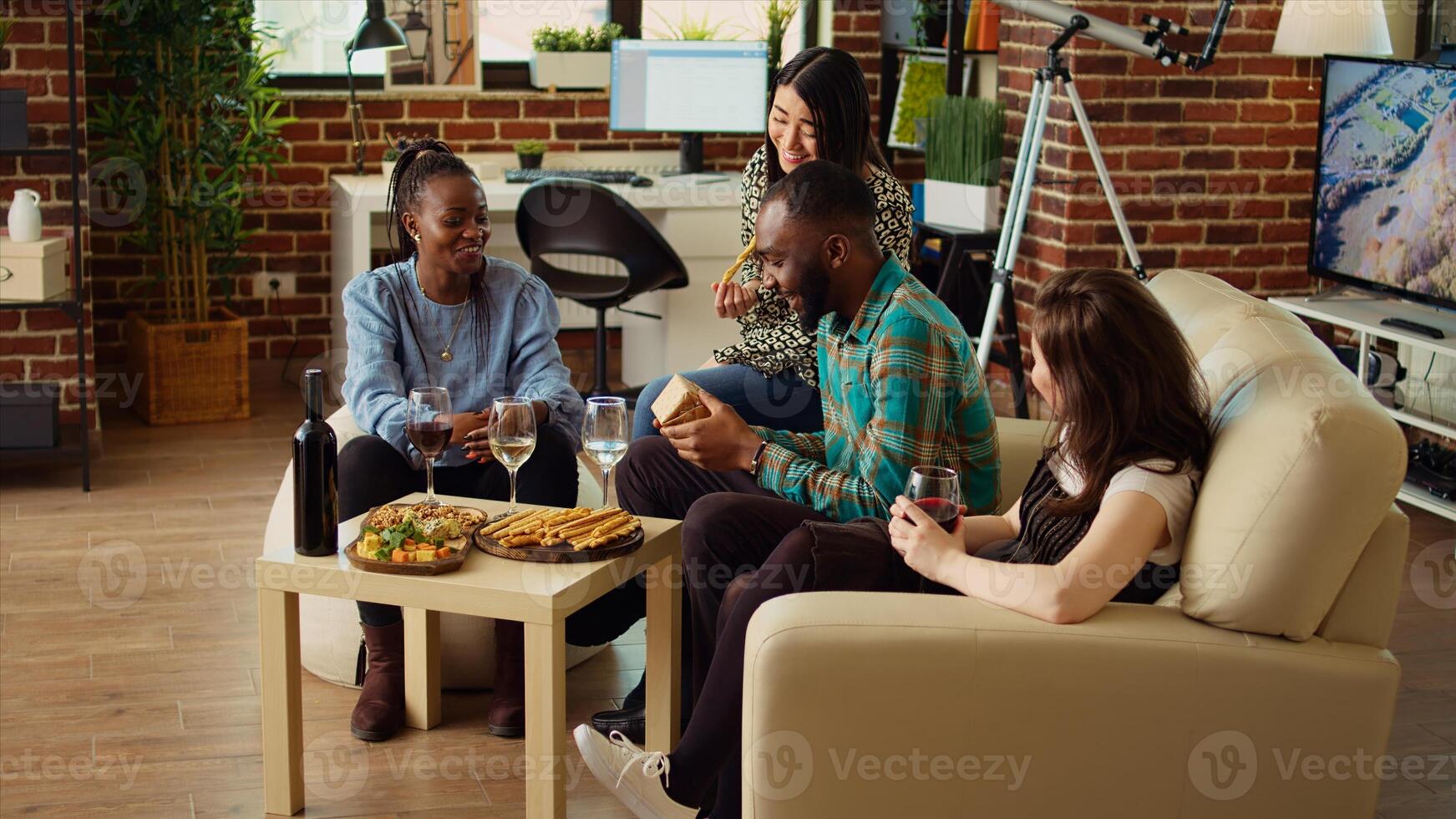 Multiethnic mates invited to birthday party offering presents to cheerful African american man impressed by nice gesture. Guests surprising their friend with thoughtful present and enjoying appetizers photo