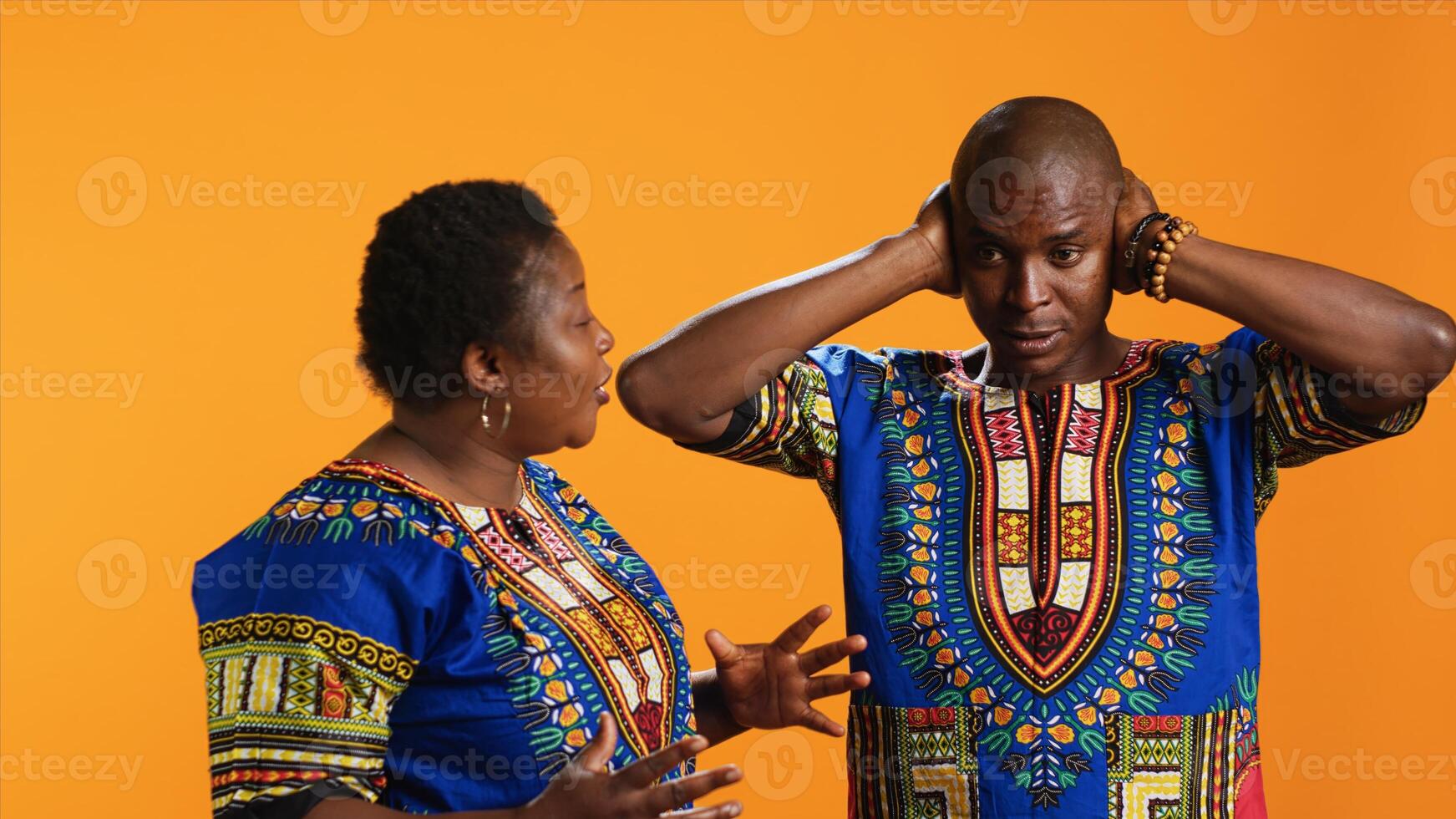 Furious girlfriend shouting at her boyfriend and fighting, having disagreement over orange background. Frustrated man covering his ears and trying to ignore his wife yelling in studio. photo
