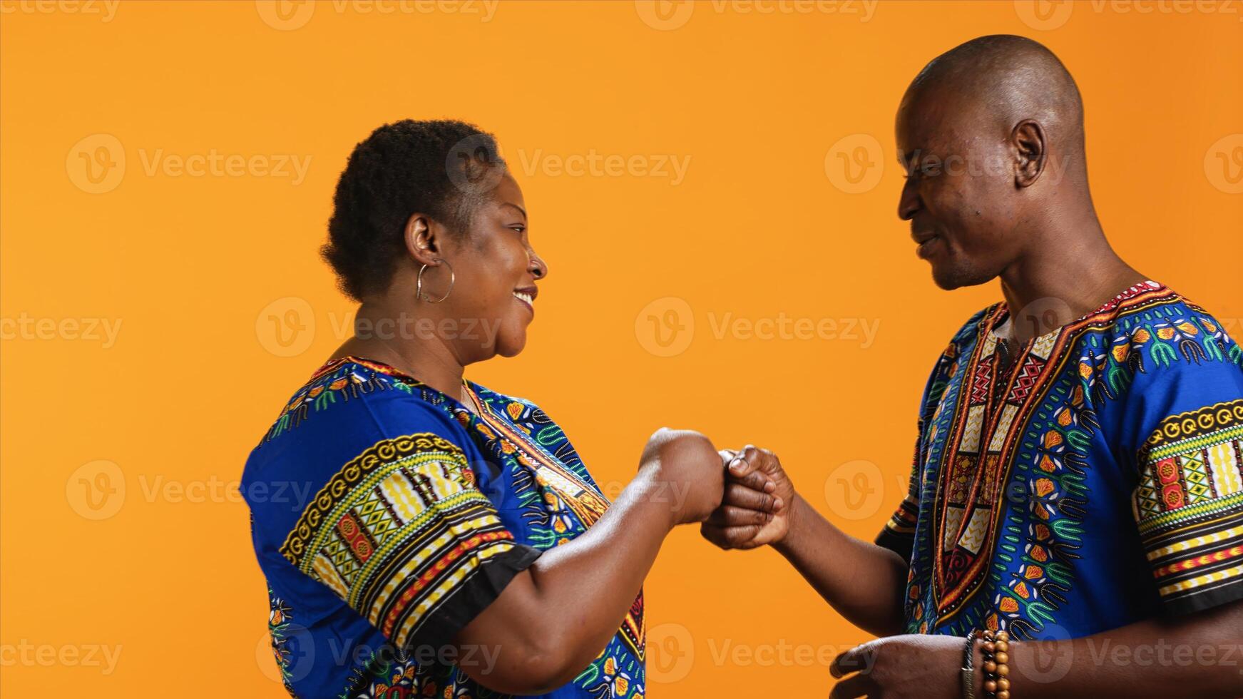 Cheerful ethnic people doing fist bump salute on camera, greeting each other with a cool modern gesture in studio. Traditional african american married couple in colorful attire having fun. photo