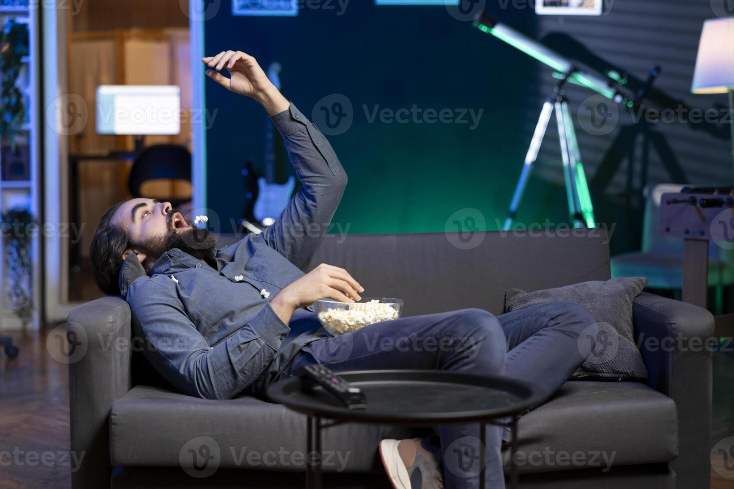 Bored man throwing popcorn in mouth, watching streaming services content on TV, having relaxing evening. Lazy viewer enjoying weekend activity in apartment, munching snacks photo