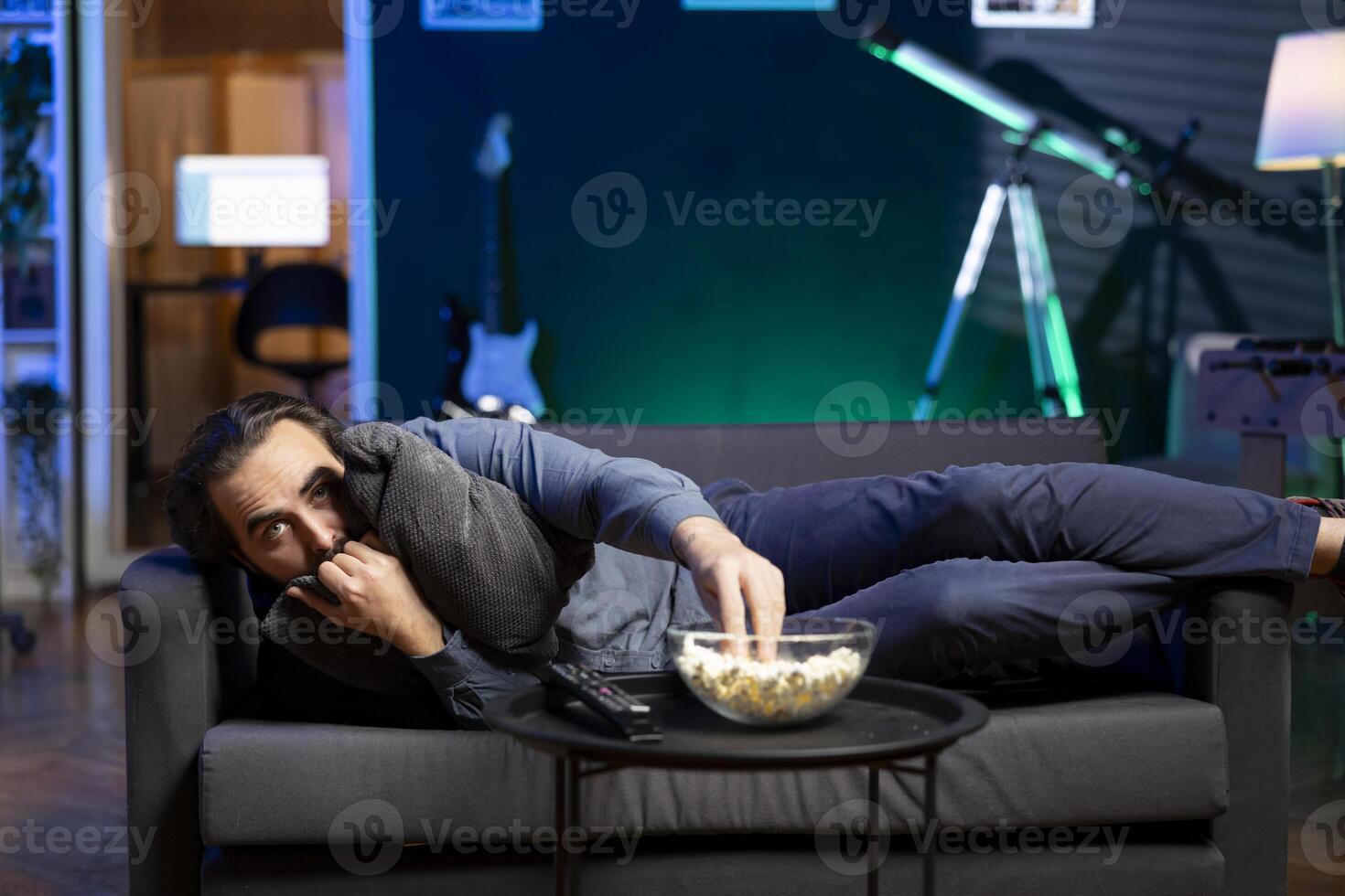 Person hugging pillow while binging suspenseful TV series on subscription based streaming services, enjoying popcorn. Scared man at home watching horror VOD shows on television display, eating snack photo